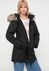 Czarna parka z kapturem wykończonym futrem, elastycznym pasem, zapięciem na zamek i bocznymi kieszeniami. Noszona na jasny sweterek w paski.