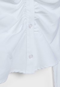 Bela tkanina z navpično vrstico gumbov, ki ima dva prosojna gumba in subtilno nabranje na straneh.