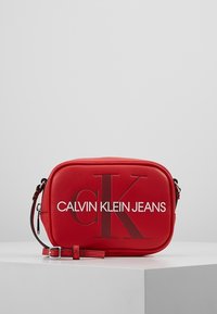 Červená obdélníková crossbody taška s nastavitelným popruhem, s vyraženým logem "cK" a bílým nápisem "Calvin Klein Jeans" na přední straně.