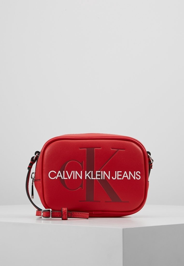 Červená obdélníková crossbody taška s nastavitelným popruhem, s vyraženým logem "cK" a bílým nápisem "Calvin Klein Jeans" na přední straně.