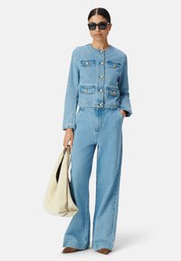 Jasnoniebieska denimowa kurtka z złotymi guzikami i kieszeniami, zestawiona z szerokimi spodniami dżinsowymi, trzymając beżową torebkę i nosząc czarne okulary przeciwsłoneczne.