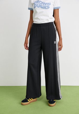Osoba nosząca czarne szerokie spodnie Adidas z białymi paskami, białą koszulkę z niebieskim wykończeniem i napisem oraz czarne buty sportowe z żółtymi paskami.