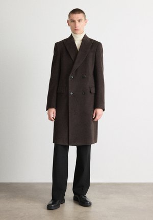 Abrigo doble faz de color marrón oscuro, hecho de tejido texturizado con botones negros, bolsillos laterales y un corte ajustado, combinado con pantalones y zapatos negros.