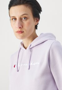 Felpa lilla realizzata in morbido tessuto. Presenta una tasca frontale e un cordino regolabile. Logo "Champion" ricamato in bianco e rosso.