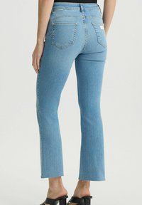 Jeans in denim azzurro chiaro con design a gamba bootcut, caratterizzati da orli sfilacciati, vita media e due tasche posteriori. Indossati con sandali neri.