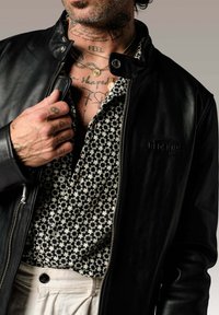 Veste bomber en cuir noir avec un col, portée par-dessus une chemise noire à motif floral. Présente des colliers superposés et des tatouages corporels visibles.