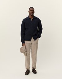 Jūras spēku adīta kardigāna ar lielām kabatām, beļģu kordura bikses, brūnas kurpes un gaismas toņa cepure, ko tur kreisajā rokā.