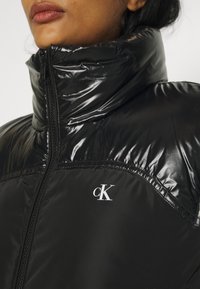 Glanzend zwarte pufferjack met hoge kraag, ritssluiting en wit "cK" logo op de borst. Gladde textuur en glanzende afwerking.