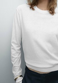 Vit sweatshirt gjord av mjukt tyg med rundad halsringning och långa ärmar, med ribbat nederkant och åtsittande manschetter. Mörka byxor syns nedanför.