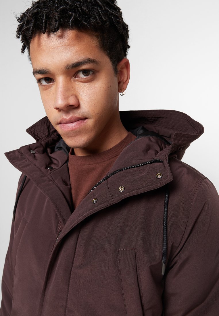 Bruine hooded jack met een ritssluiting aan de voorkant, drukknoopjes en trekkoorden. Heeft een textuurstof en een borstzak.