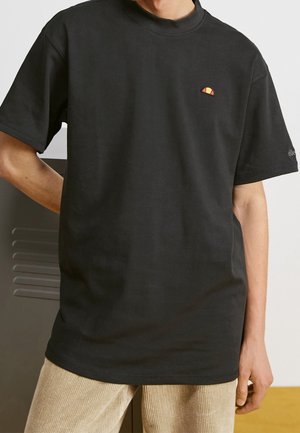 Person iført en plain sort t-shirt med korte ærmer, med en lille orange og gul logo på brystet og beige bukser.