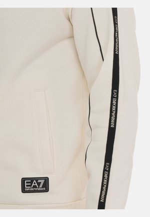 Krämmfärgad sweatshirt med svarta randiga detaljer på ärmarna. Har en patch med "EA7 Emporio Armani" och sidofickor. Mjuk textur.