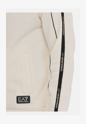 Sudadera color crema con acentos en negro a rayas en las mangas. Presenta una etiqueta de parche con "EA7 Emporio Armani" y bolsillos laterales. Textura suave.