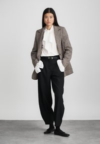 Ruskea houndstooth-bleiseri valkoisen, rusetilla varustetun paidan päällä, mustat korkeavyötäröiset housut ja mustat kengät luovat räätälöidyn ilmeen.
