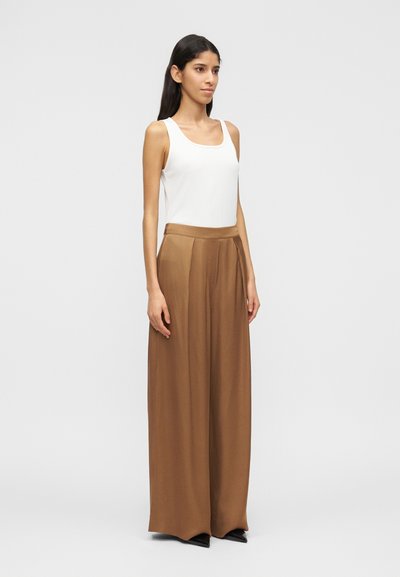 Γυναίκα όρθια με τα χέρια στα πλάγια, φοράει λευκό αμάνικο τοπ, καφέ φαρδιά παντελ όνα με ψηλή μέση και μαύρα μυτερά παπούτσια, σε απλό φόντο.