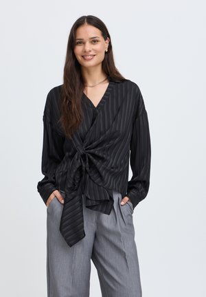 Jeune femme aux longs cheveux foncés sourit, portant un chemisier cache-cœur rayé noir et un pantalon gris à jambes larges, debout les mains dans les poches.