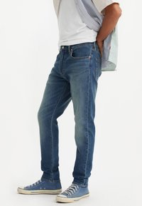 Jeans de mezclilla azul de corte recto, con un diseño clásico de cinco bolsillos y costuras naranjas, combinados con zapatillas de lona azules.