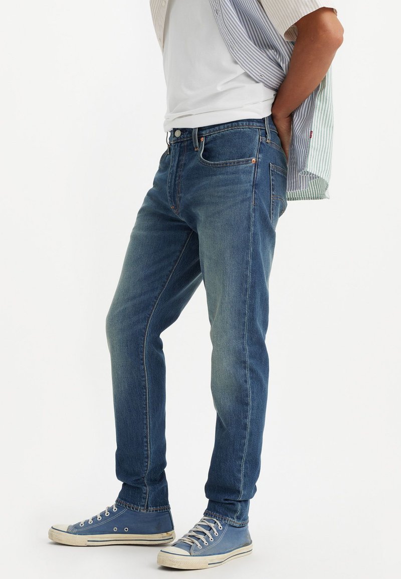 Blå denimjeans med rak passform, med en klassisk femfickdesign och orange sömmar, i kombination med blå kanvas sneakers.