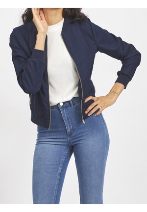 Kobieta ubrana w granatową kurtkę na zamek na białej bluzce oraz niebieskie, wysokie dżinsy, pozująca z jedną ręką na biodrze, a drugą przy twarzy.