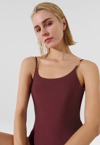 Burgundské plavky s hladkým povrchem, výstřihem ve tvaru lodi a tenkými ramínky. Na ramínku je zlatý detail pro zvýraznění designu.