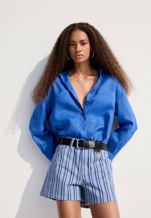 Mujer con pelo largo y rizado que lleva una camisa azul abotonada metida en unos pantalones cortos a rayas azules y blancas con un cinturón negro, de pie contra una pared blanca.