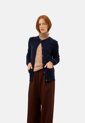 Marineblå cardigan i blødt strik med rund halsudskæring, knaplukning foran og to forlommer, lagdelt over en beige turtleneck og brune bukser.
