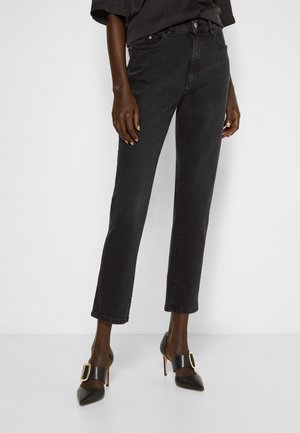 Kobieta ubrana w czarne dżinsy z wysokim stanem i prostymi nogawkami oraz czarne szpilki z paskami na złote klamry, stojąca na białym tle.