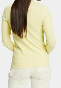 Femme aux longs cheveux bruns portant une chemise à manches longues jaune pâle et un pantalon blanc cassé, vue de dos sur fond blanc.