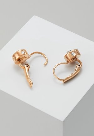 Paire de boucles d'oreilles clips dorées avec des dessus arrondis comportant de petites pierres précieuses transparentes, présentées sur un cube blanc sur fond gris.