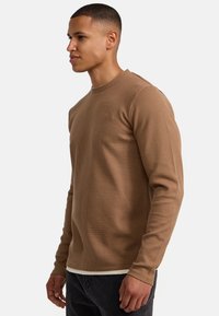 Braunes langärmliges Shirt mit Rippenmuster und rundem Ausschnitt aus strukturiertem Stoff, über ein weißes Unterhemd getragen, kombiniert mit dunklen Jeans.