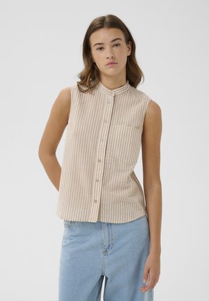 Młoda kobieta w beżowo-białej paskowej bezrękawnikowej koszuli zapinanej na guziki i jasnoniebieskich dżinsach z wysokim stanem na jednolitym tle.