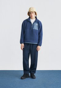 Νεαρός άνδρας που φοράει μπεζ σκουφάκι από φλις με προστασία αυτιών, μπλε σκούρο πουλόβερ φλις με τσέπη στο στήθος, παντελόνι σετ και μαύρα αθλητικά παπούτσια, όρθιος ακίνητος.