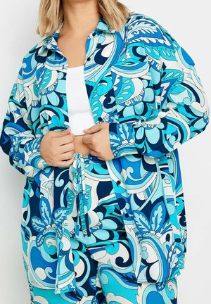 Femme portant une surchemise et un pantalon assortis bleu et bleu sarcelle à motifs, associés à un débardeur court blanc, mains ajustant la chemise.