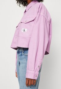 Lila denimjacka i kort modell med krage, två framfickor och en logopatch. Har knäppning vid ärmsluten och en avslappnad passform.