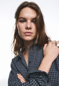 Košeľa s dlhými rukávmi na gombíky, zdobená námorníckym modrým a bielym kockovaným vzorom. Tkanina pôsobí mäkko, s voľným strihom a vyhrnutými rukávmi.