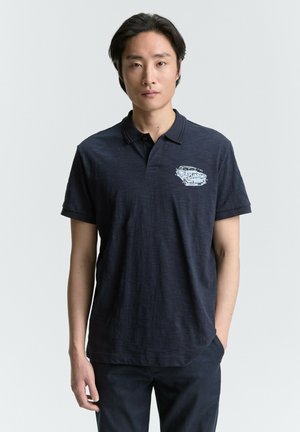 Polo shirt navy realizzato in tessuto testurizzato, con colletto corto, maniche corte e una grafica stampata sul lato sinistro del petto.