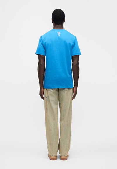 Μπλε βαμβακερό μπλουζάκι με κοντά μανίκια, με μικρό λευκό γραφικό αστροναύτη στην πλάτη, συνδυασμένο με ανοιχτό μπεζ παντελόνι φαρδιάς γραμμής.