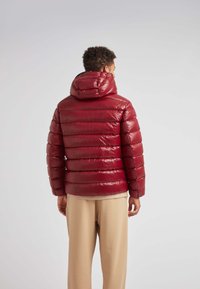 Rød puffjakke med blank finish, quiltet design og hætte. Båret over beige bukser. Bagperspektiv viser vandret syning.
