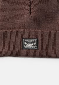 Gorro de malha canelada castanho com um rótulo preto apresentando texto branco e um gráfico de cavalos. Textura suave com um ajuste justo.