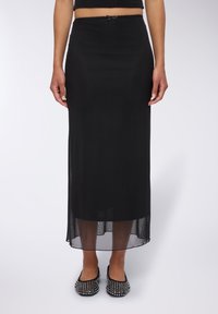 Gonna nera maxi con sovrapposizione trasparente e vita elastica, caratterizzata da un piccolo dettaglio a fiocco. Indossata con ballerine nere decorate.