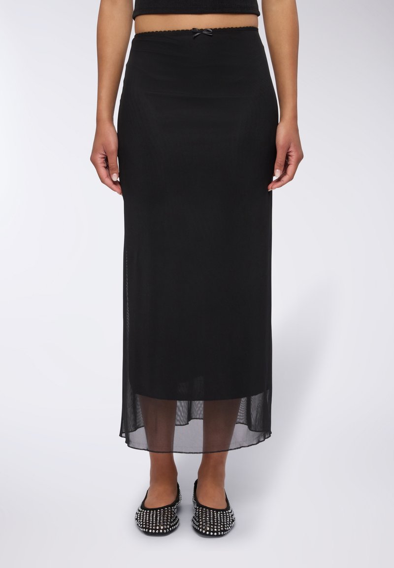 Gonna nera maxi con sovrapposizione trasparente e vita elastica, caratterizzata da un piccolo dettaglio a fiocco. Indossata con ballerine nere decorate.