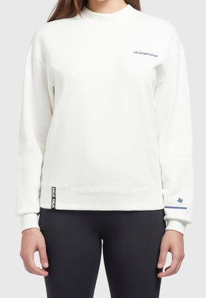 Kobieta w jednolitej białej bluzie z małym granatowym napisem na piersi i wzorem liścia klonu przy lewym nadgarstku, w połączeniu z czarnymi legginsami.