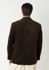 Blazer en velours côtelé marron avec une surface texturée, une fente unique à l'arrière, et des boutons décoratifs sur les manches, affichant une coupe ajustée.