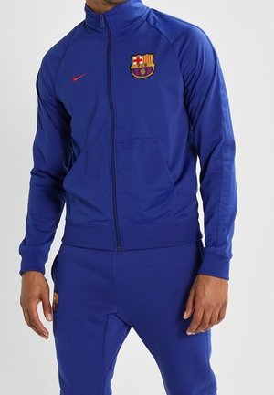 Niebieski dres na całej długości suwaka z herbem FC Barcelona i czerwonym logo Nike noszony przez osobę, pokazującą kurtkę i pasujące spodnie.