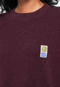 Bordový sveter s textúrovanou látkou, s farebným okrúhlym logom, na ktorom je napísané "HOUSE of COLORS." Klasický dizajn s okrúhlym výstrihom.