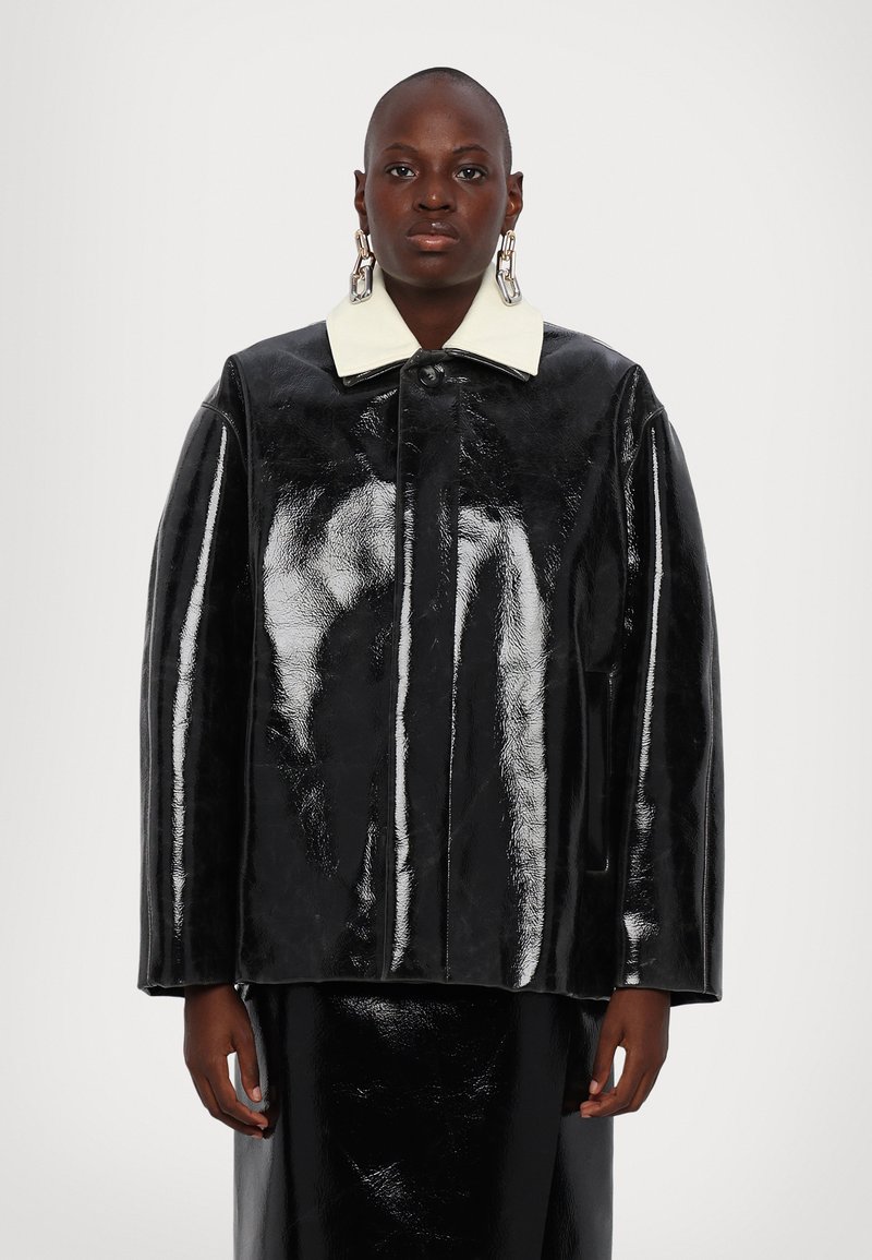 Giacca in pelle verniciata nera con colletto bianco, vestibilità oversize, chiusura frontale a bottoni e superficie liscia e lucida.