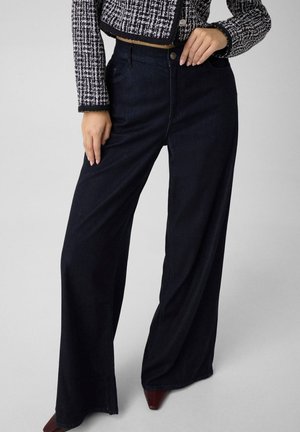 Femme portant un pantalon noir taille haute à jambes larges, une veste en tweed noir et blanc et des chaussures pointues marron foncé, debout avec une main sur la taille.