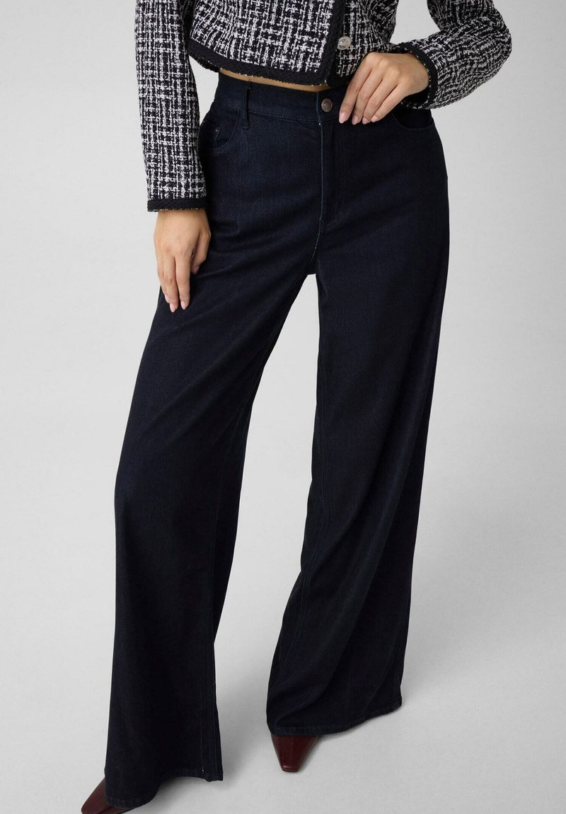 Femme portant un pantalon noir taille haute à jambes larges, une veste en tweed noir et blanc et des chaussures pointues marron foncé, debout avec une main sur la taille.