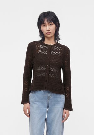 Ung kvinde iført en mørkebrun hæklede cardigan med knapper og lyseblå jeans, der står mod en ensfarvet hvid baggrund.