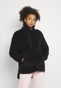 Pull en polaire noir avec col montant, fermeture éclair à mi-hauteur et poches avant. Texture douce et coupe décontractée, avec un dos plus long.